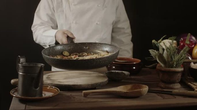 穿着白色制服的男厨师拿着煎锅，用新鲜蔬菜炒意大利面