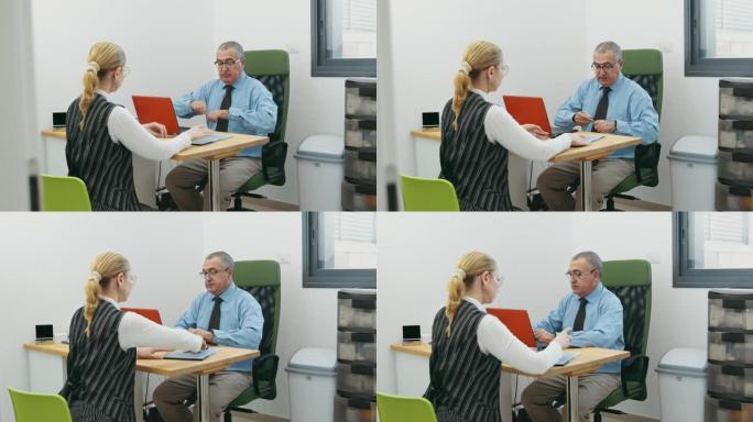 商人和办公室里的女人在桌子旁用笔记本电脑交谈，在咨询室里填写文件