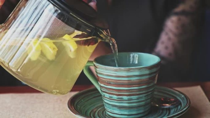 Lass从透明的玻璃水壶中倒入热凉茶，蒸香草，薄荷和几片柠檬漂浮在条纹陶瓷杯中。