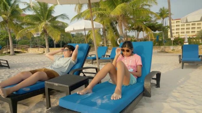 快乐的亚洲儿童戴着墨镜，早上在酒店或度假胜地度假时一起在沙滩椅上放松，并带有家庭生活方式的概念。