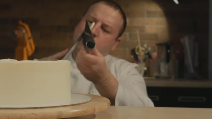 一位高加索男性糕点厨师坐在膝盖上，将完美的蛋糕空白对齐。去除多余的奶油，用糕点刮刀刷掉。专注和专业