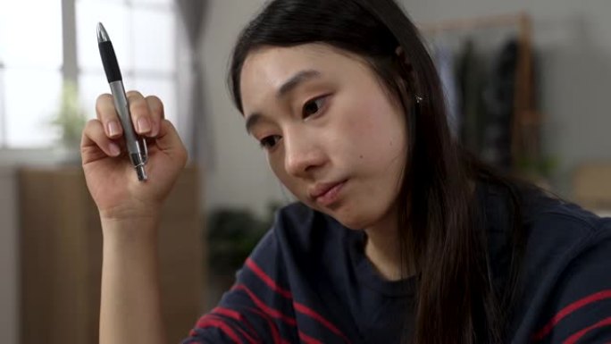 一个无聊的亚洲女大学生在家学习的特写镜头正在旋转笔，凝视着空旷的地方，同时在卧室里上在线课程。