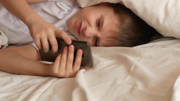 早上7-9岁的高加索男孩在床上关闭智能手机上的闹钟，用枕头遮住耳朵，继续睡觉。这个男生不想醒来上学。