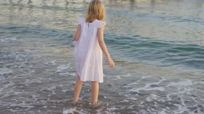 一个穿着白色连衣裙的可爱小女孩赤脚走进冰冷的海水。