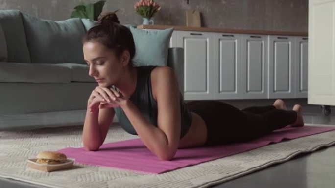 一个女人躺在健身床垫上的视频，双手放在下巴下，看着她面前的汉堡。健康的生活方式