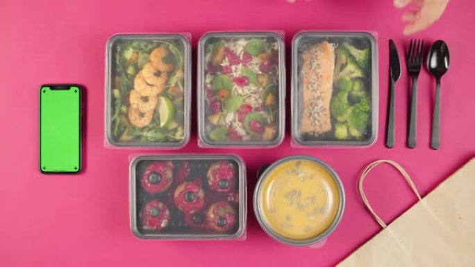 送餐顶视图，在粉红色背景的一次性容器中带走餐点。午餐盒配有煮熟的素食菜肴，使用带色度绿屏的手机。健康