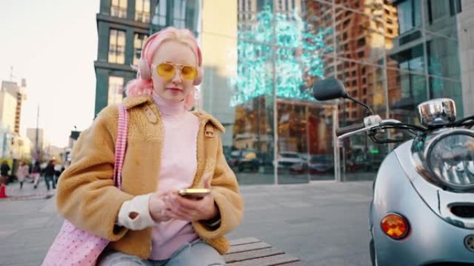 年轻时尚的女人，留着粉红色的发型，坐在户外，戴着大大的无线耳机，听着音乐