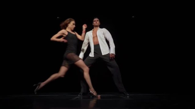 交际舞情侣在黑暗的舞台上，舞蹈表演部分。身穿黑色透明短裙的女子跑到身穿白色衬衫未扣的男子面前，跳了起