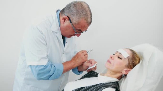 整形外科医生做两次填充注射透明质酸在鼻唇沟妇女病人的脸躺在椅子上。患者注射后感到疼痛