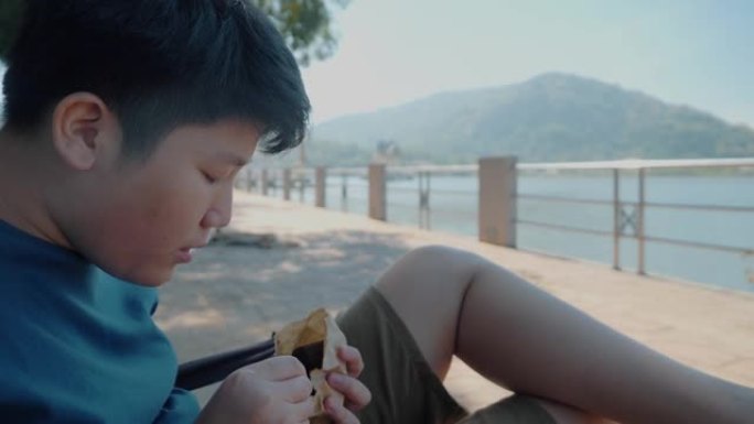 快乐的亚洲男孩在阳光明媚的日子里吃一些零食，看自然背景的水景，生活方式的概念。