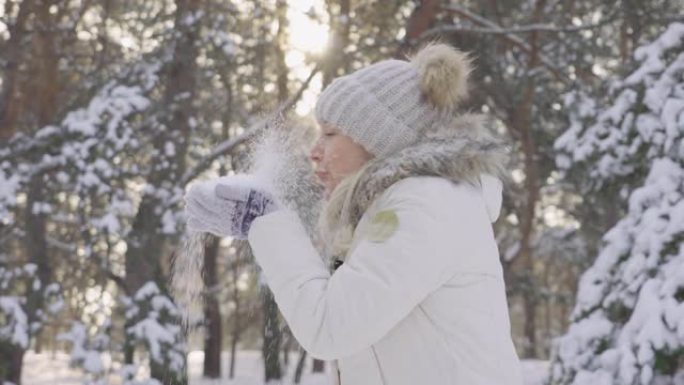 侧面可爱的女孩穿着暖和的冬衣，把雪抛起来，开心地笑。美丽的模糊背景雪域自然松林。缓慢的运动。近距离