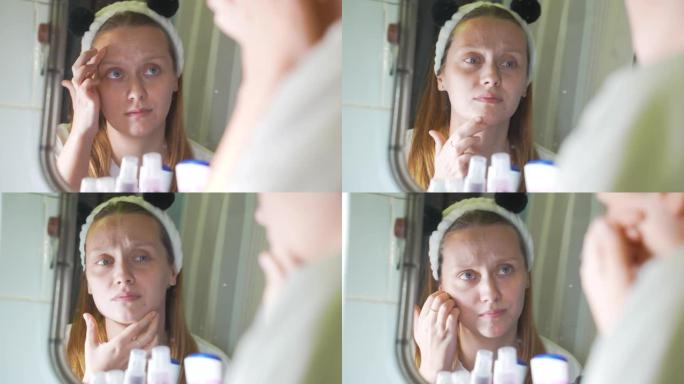 年轻的白人妇女在浴室的镜子里看着自己，检查她的脸部皮肤，感到不安。脸上有红肿和丘疹。这个女孩很沮丧。