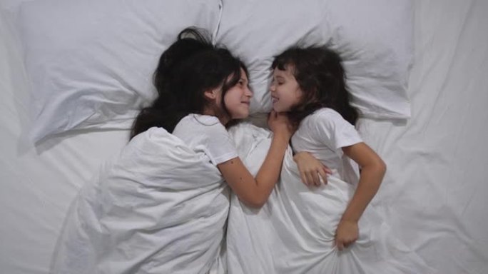 高加索人的俯视图两个孩子在床上用白色床单吵架。高角度拍摄。小姐妹争辩，互相冒犯，然后转向不同的方向