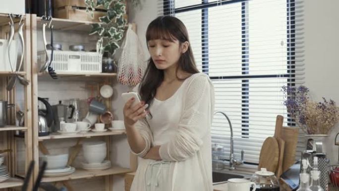 昏昏欲睡的亚洲女性穿着休闲服靠在厨房水槽上，正在打哈欠，一边喝着早茶，一边在家用智能手机浏览在线新闻