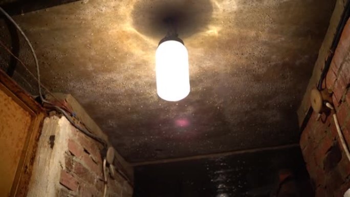 一盏旧的肮脏的圆柱形灯挂在砖墙上的深色地下室电线中的混凝土天花板上