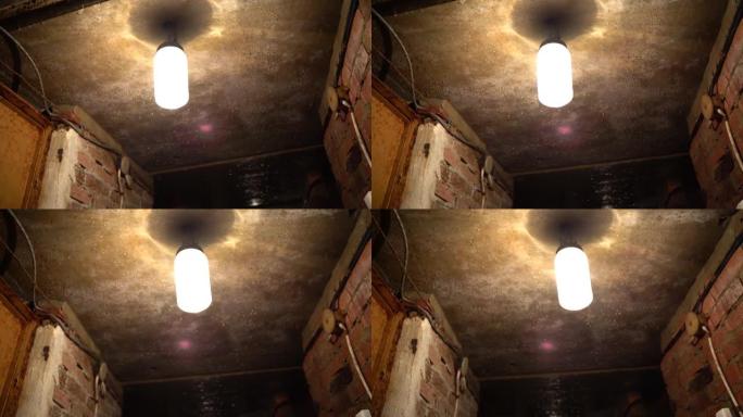一盏旧的肮脏的圆柱形灯挂在砖墙上的深色地下室电线中的混凝土天花板上