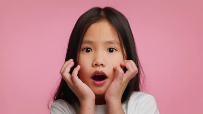惊讶的韩国小女孩触摸脸在休克，粉红色背景
