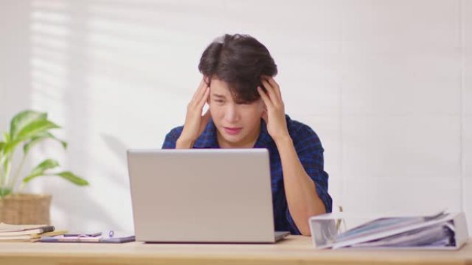 压力很大的亚洲商人对他的工作有问题，所以头痛和偏头痛。在家里工作的桌子上受罪的年轻人。压力男人的概念