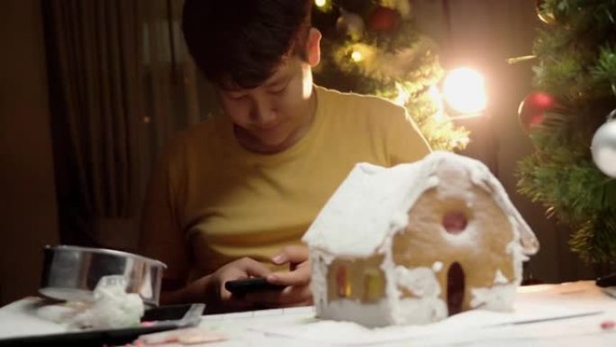 亚洲男孩在自制姜饼屋附近使用智能手机，圣诞节庆祝生活方式概念。