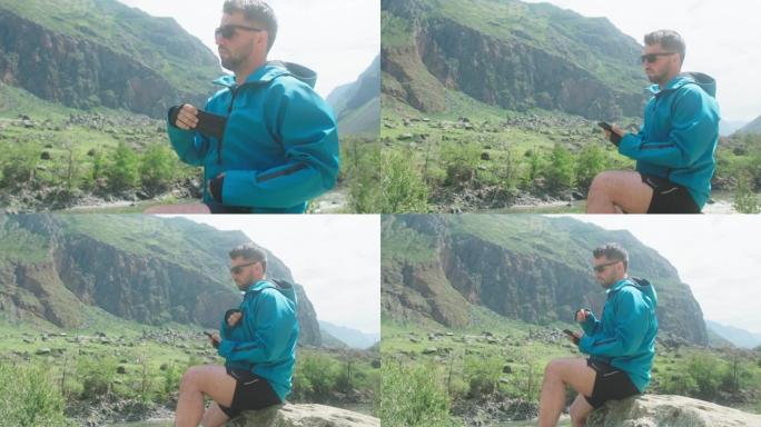 一个戴着眼镜的男人坐在悬崖边，戴着蓝色外套，掏出手机。卡图-雅里克峡谷丘里什曼山谷。阿尔泰