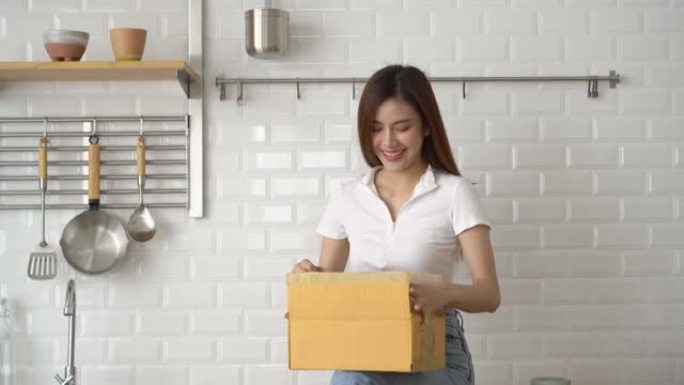 美丽的亚洲女人坐在时尚厨房的柜台上，打开包装有在线订单的纸板包装，感觉很开心。兴奋的女孩在网上打开盒