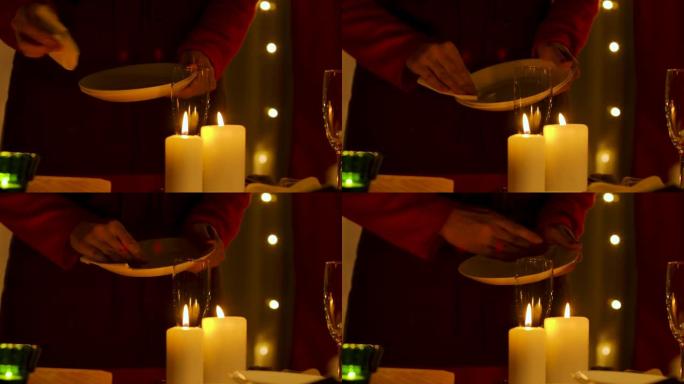 女人在装饰着庆祝新年或圣诞节的房间的背景下擦拭白色盘子。蜡烛在眼镜旁边的节日桌子上燃烧。光线不足。双