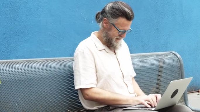 中年男子坐在街上的长椅上在笔记本电脑上打字。