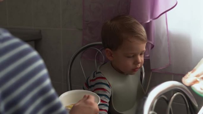 食物，营养，童年概念-中等射击妈妈用橡胶勺子喂养婴儿男孩。可爱的饥饿的孩子穿着围兜坐在孩子们的椅子上