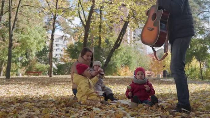 年轻的母亲保姆在公园里野餐，三个小孩坐在毯子上。孩子们在秋天公园的篮子寒冷天气里吃糖果糖果黄色落叶。