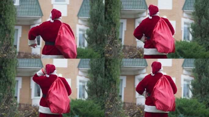后视图圣诞老人带着红色的袋子走到家里挠头思考。圣诞老人穿着红色服装，后院户外有礼物。新年惊喜和庆祝概