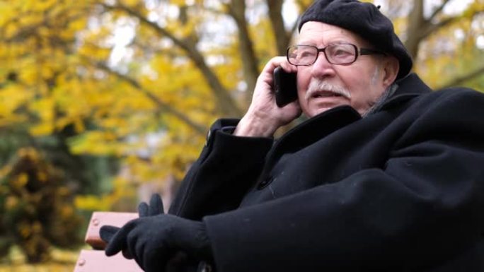 积极的老人放松，秋天的城市10月公园坐在长凳上叫第二个智能手机。
