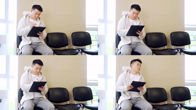 年轻的亚裔男子学生坐在候诊室的椅子上，填写申请表。男性在医院接待或办公室工作