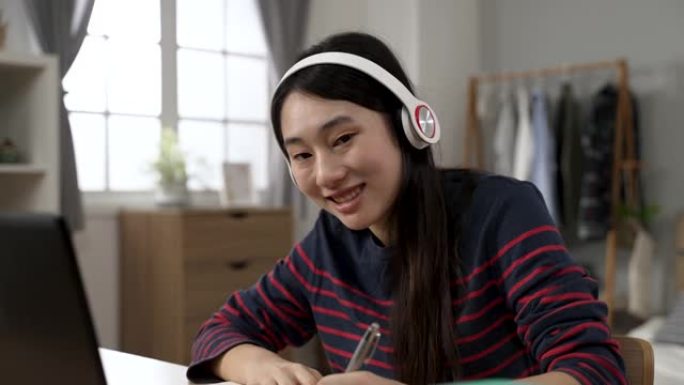 一位亚洲女大学生用耳机写作业的肖像在白天在家学习时转向镜头并微笑。