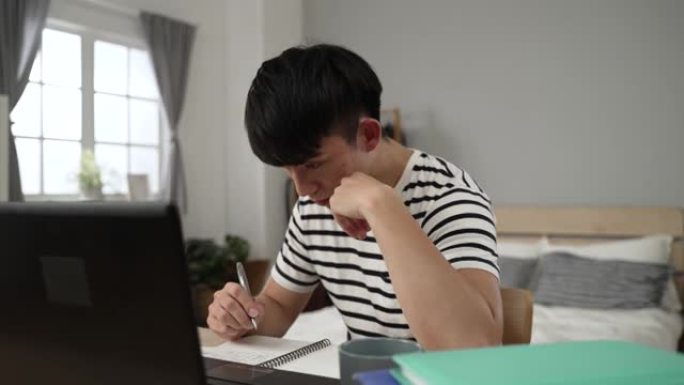疲惫的亚洲大学生从家里学习，从打瞌睡中醒来，正试图在卧室用电脑写作业的同时专注于学习。