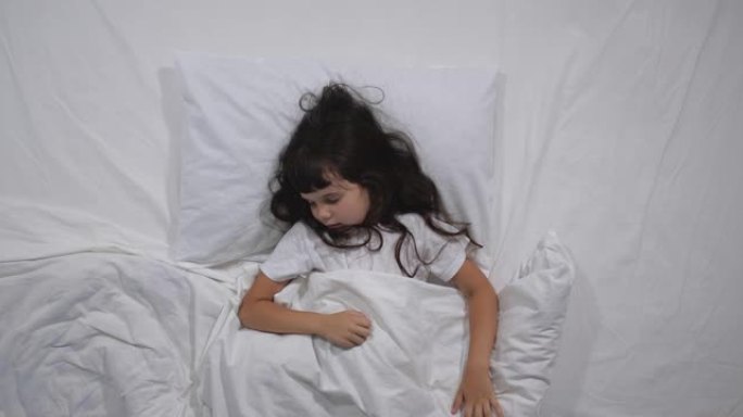6岁的小女孩的俯视图，长长的棕色头发在床上睡着了，白色床单在明亮的房间里。高角度拍摄。学龄前女孩要睡