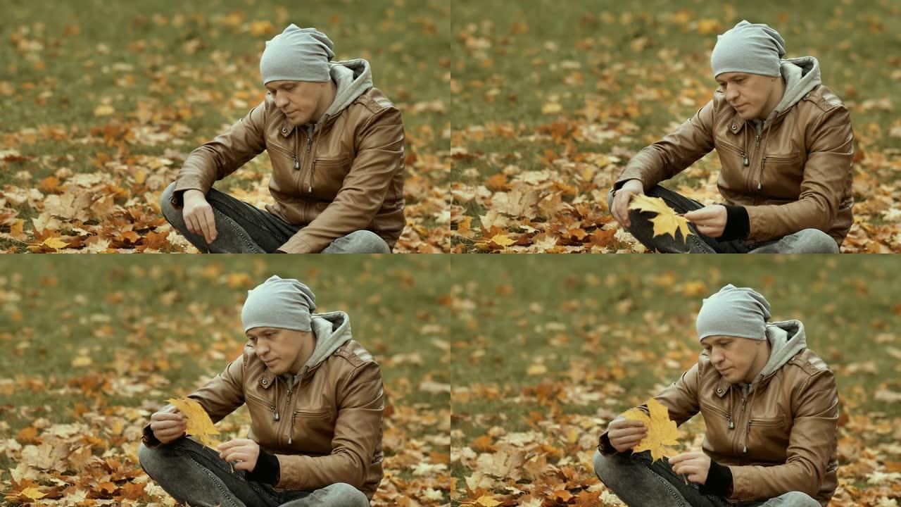 一个戴着帽子的悲伤的人坐在落叶之间，看着一片泛黄的枫叶，秋天的忧郁