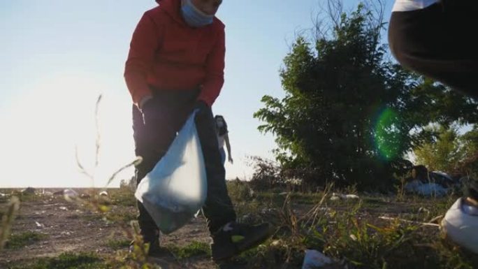 靠近生态活动家的家庭，戴着口罩清洁农村废纸草坪。年轻的父母和戴着手套的孩子在路边附近的袋子里收集垃圾