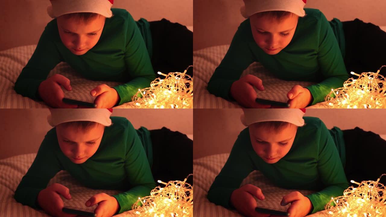 一个晚上躺在床上的圣诞装饰品的男孩点击智能手机屏幕，看着屏幕。