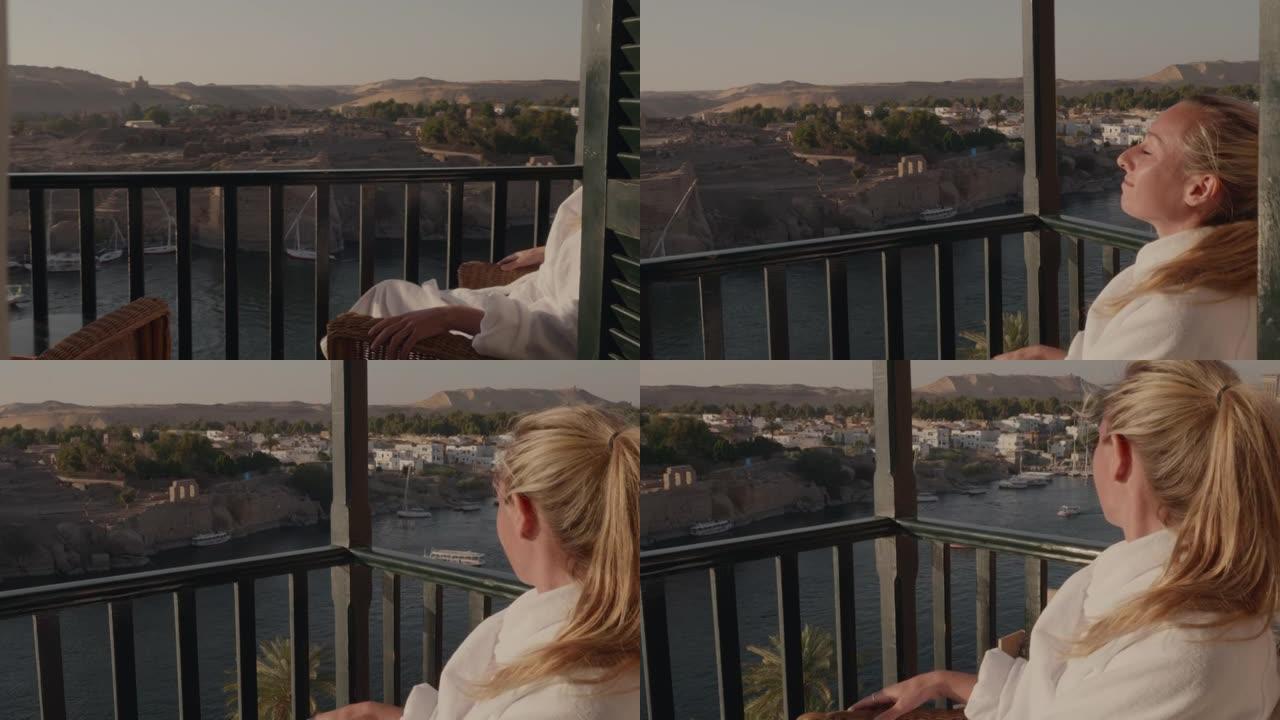 年轻女子在酒店的阳台上穿着浴袍，在埃及阿斯旺 (Aswan) 欣赏壮丽的河景。女性旅行需要一点时间来