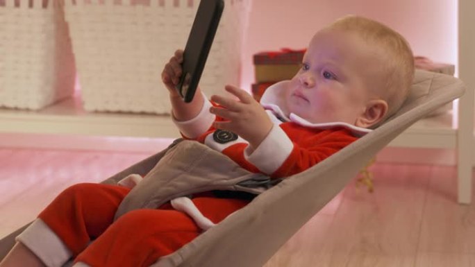 快乐的婴儿穿着圣诞老人服装，使用智能手机坐在房间里，带着圣诞节日的灯光，可爱的孩子庆祝他的第一个新年