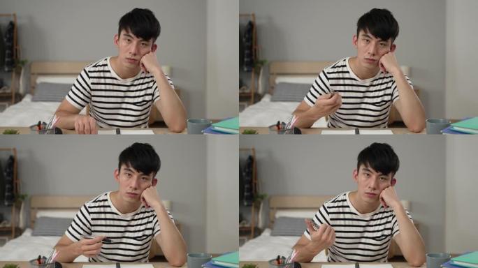 无聊的亚洲男学生盯着相机在家里学习时托着头玩笔在卧室看无聊的在线讲座视频