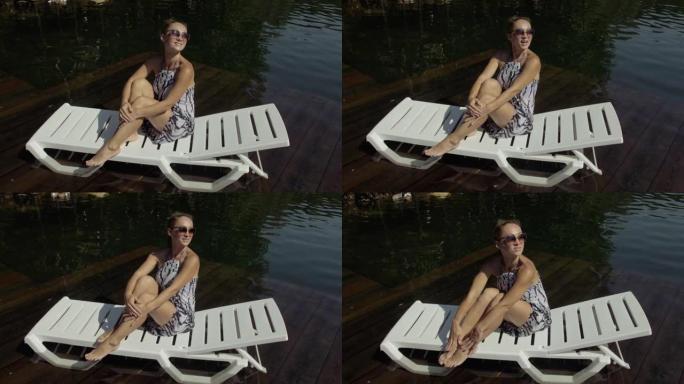 女人躺在太阳镜和波西米亚丝绸披肩的日光浴床上。女孩在洪水木水下码头上休息。湖里铺满了水