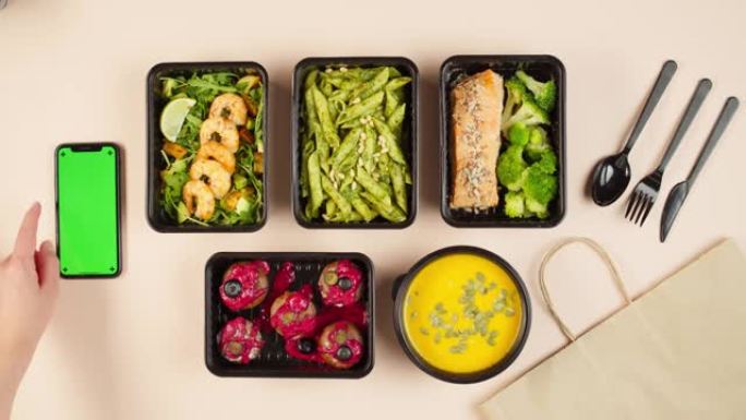 送餐俯视图，用一次性容器带走餐点。午餐盒配有煮熟的素食菜肴，使用带色度绿屏的手机。健康饮食。餐饮服务