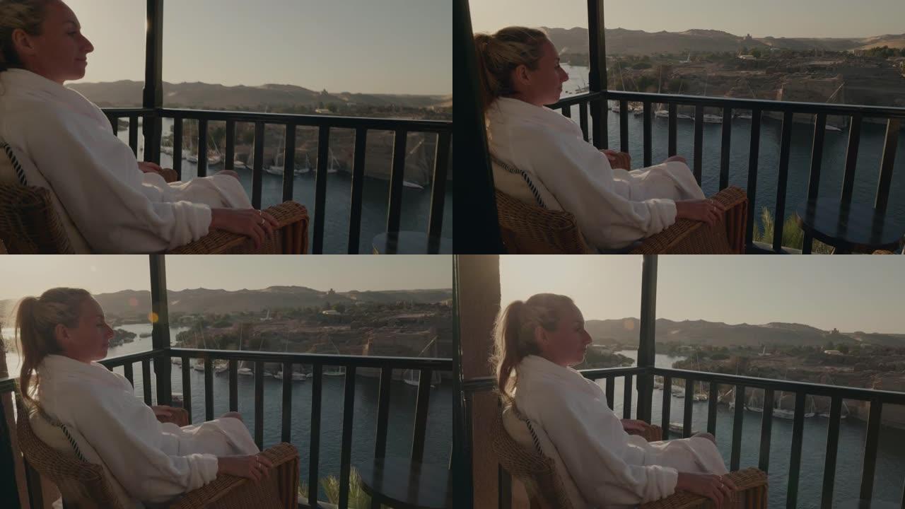 年轻女子在酒店的阳台上穿着浴袍，在埃及阿斯旺 (Aswan) 欣赏壮丽的河景。女性旅行需要一点时间来