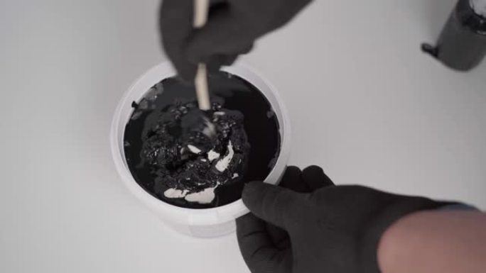 画家将黑色的油漆倒入带有白色丙烯酸腻子的桶中，并用棍子搅拌混合物