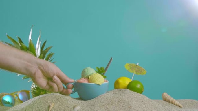 用手在杯子里拿起冰淇淋，旁边是沙子里的菠萝和柠檬装饰品。