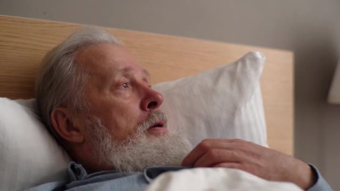 生病的白发成熟成年男性患者的特写脸躺在床上，向疗养院的医生说些什么。
