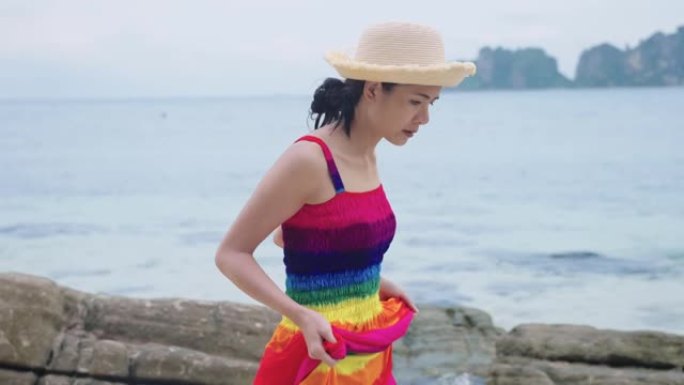 侧视图一位年轻迷人的亚洲女性举起彩虹裙，享受在海滩岩石海岸线、热带岩石海岸、赤脚在海滩探索、阳光明媚