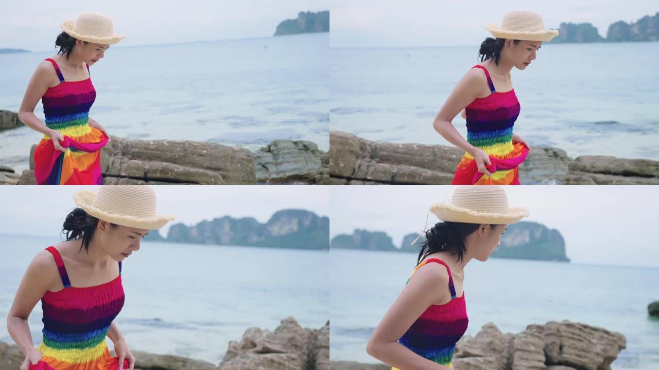 侧视图一位年轻迷人的亚洲女性举起彩虹裙，享受在海滩岩石海岸线、热带岩石海岸、赤脚在海滩探索、阳光明媚