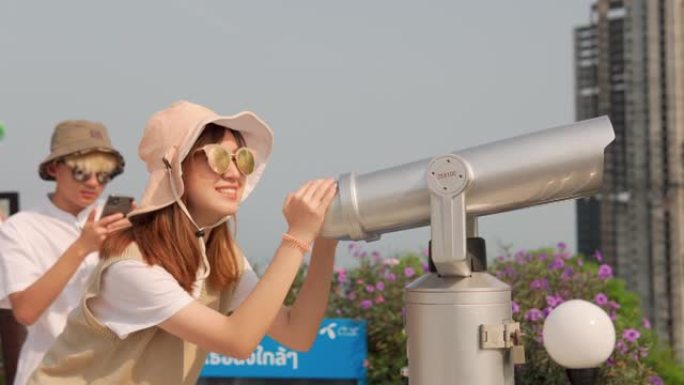 欢快的亚洲年轻夫妇游客在观看点使用投币式双筒望远镜观看城市背景。
